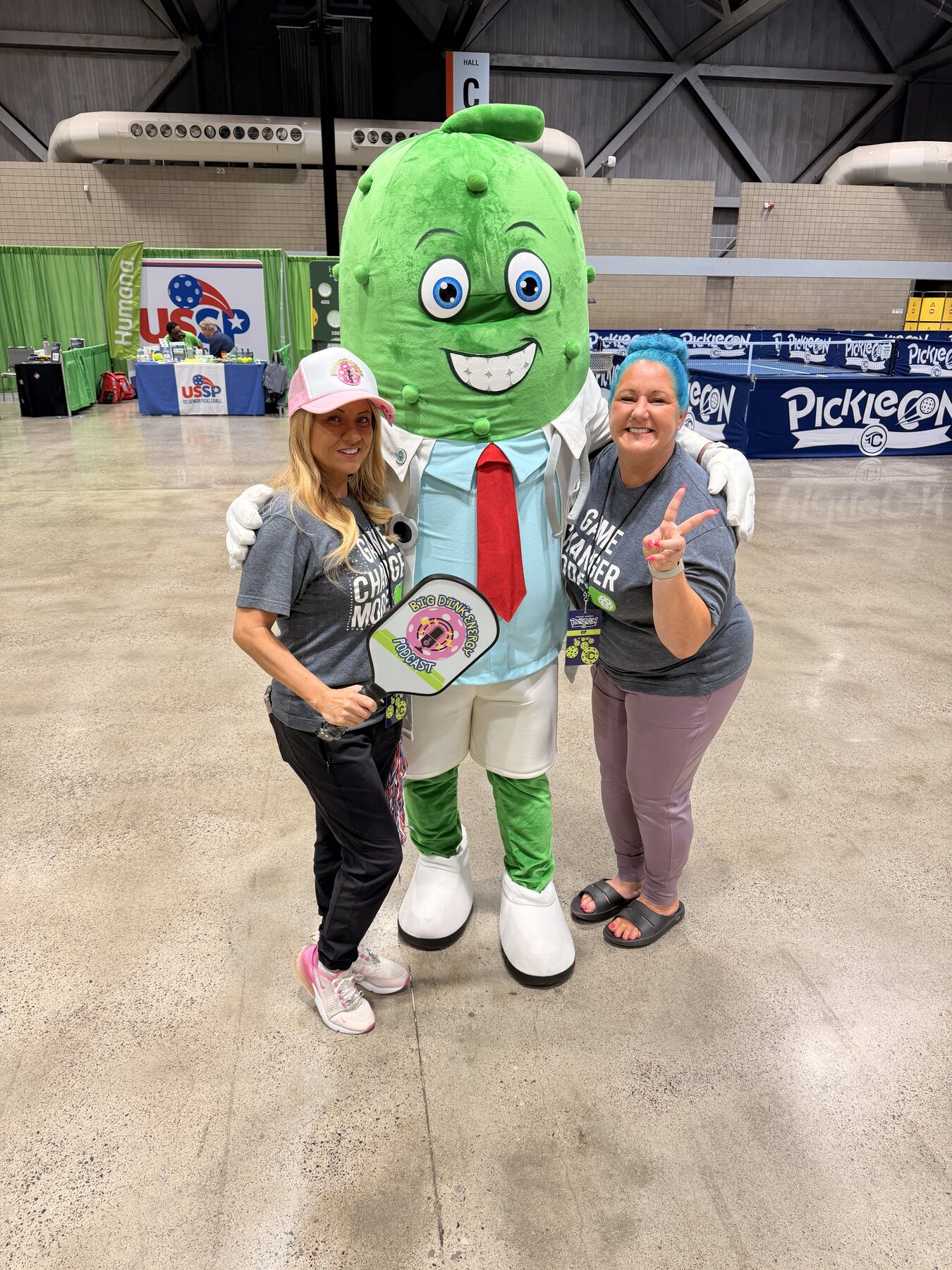 Two event attendees posing beside a pickleball mascot at PickleCon